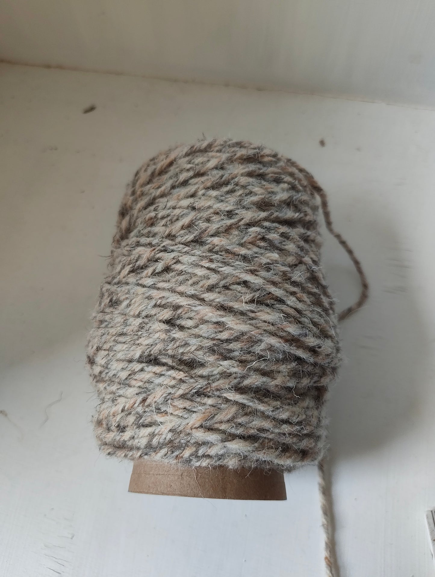 A spool of thick, twisted weaving yarn in natural brown, grey and beige shades, wound on a brown cardboard core against a white background.
