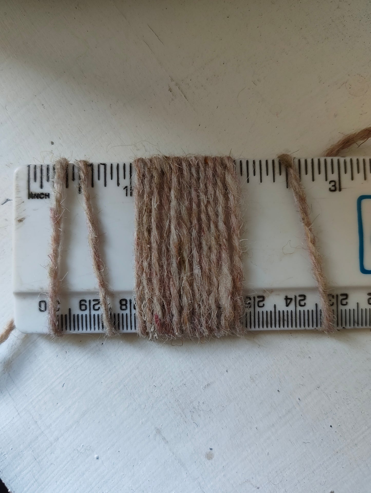 A close-up of a beige and brown yarn or thread swatch wrapped around a white plastic ruler to show texture and color, placed on a white surface.