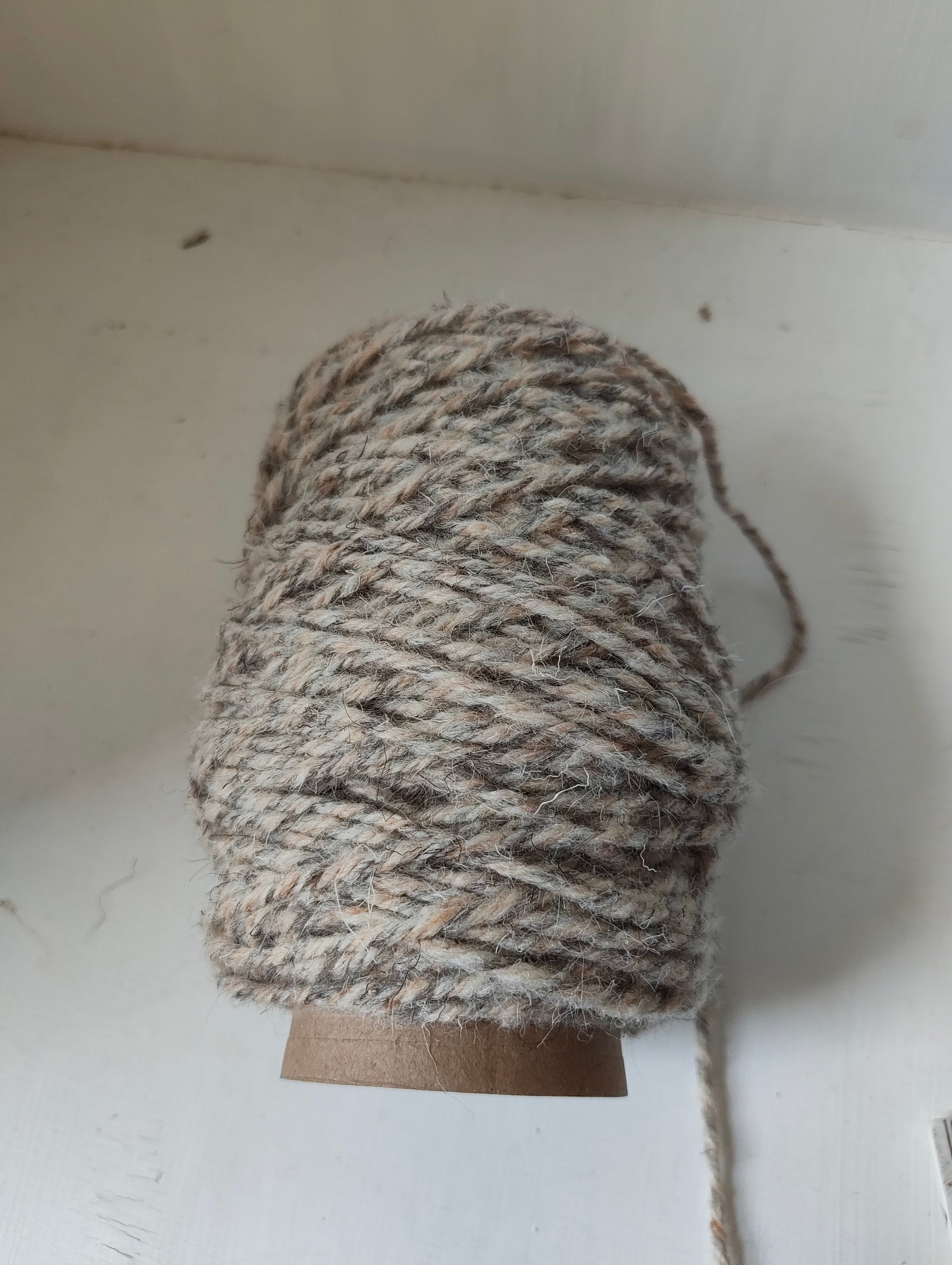 A spool of thick, twisted weaving yarn in natural brown, grey and beige shades, wound on a brown cardboard core against a white background.
