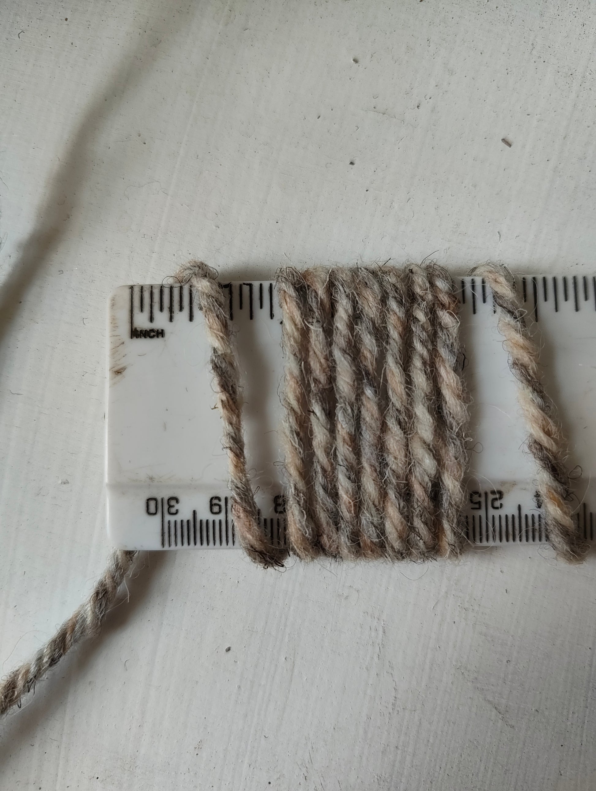 A close-up of thick, twisted wool yarn in cream, sand and grey shades wrapped around a white plastic ruler to show its texture and thickness, set against a light off-white background.