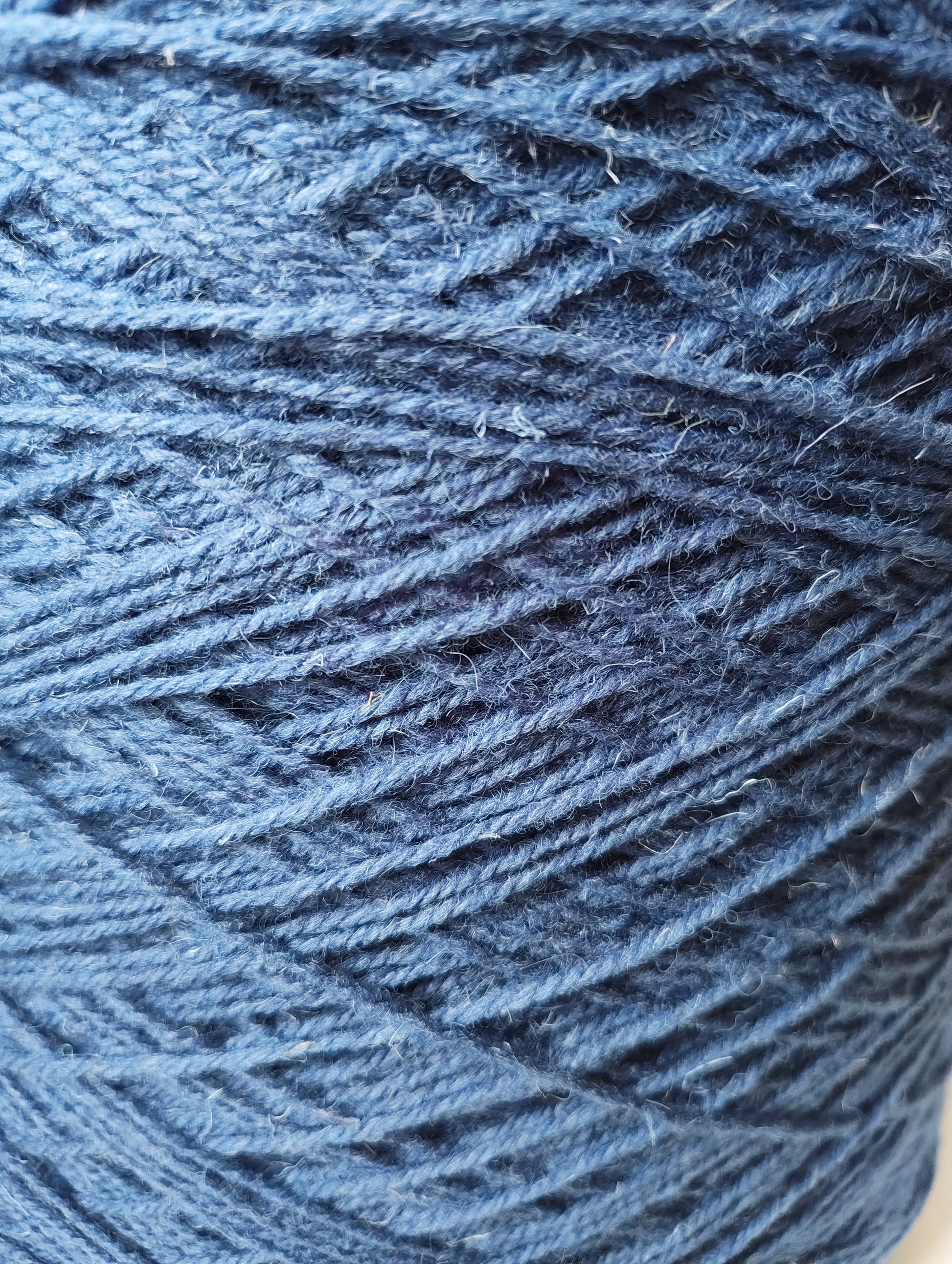 A close-up of a medium blue weaving yarn wound into a cone, showing the textured, fibrous strands of the rug yarn.